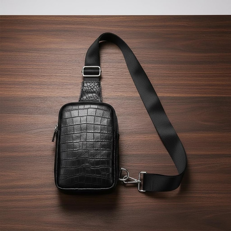 May include: Black crossbody bag with a textured, crocodile-like pattern. The bag has a zippered main compartment and a long, adjustable black strap. The bag is made of leather and has silver-tone hardware. The bag is shown on a dark wood surface.