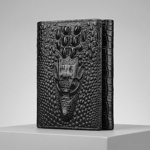 Può includere: Un portafoglio in pelle nera caratterizzato da un prominente design tridimensionale in rilievo a forma di testa di coccodrillo sulla copertina anteriore. La superficie presenta una texture a scaglie, con una finitura lucida e orientamento verticale.