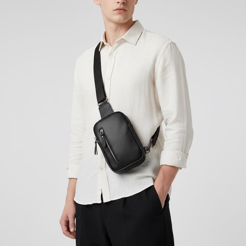 May include: Black crossbody bag with a textured surface and a zippered front pocket. The bag has a black adjustable strap and a silver-tone metal clasp. The bag is worn over the shoulder.
