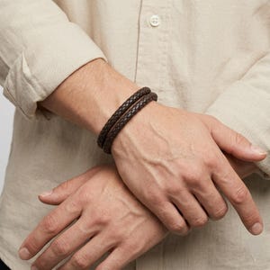 May include: Two dark brown braided leather bracelets. The bracelets are worn on a person's wrist. The bracelets are made of a woven leather material. The person is wearing a light tan button-down shirt.