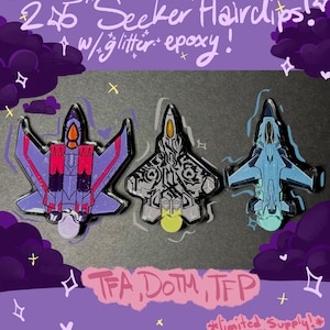 May include: Three 2.5 inch Seeker hairclips with glitter epoxy. The left clip is purple and red, the center clip is black and white with intricate patterns, and the right clip is light blue. Text reads: 2.5" Seeker Hairclips! w/ glitter epoxy! TFA, DOTM, TFP, limited supply!