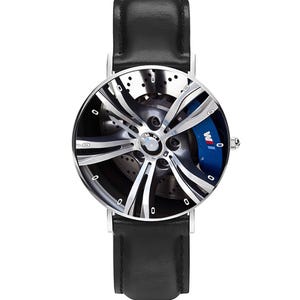 May include: A wristwatch with a black leather band and a silver-toned watch face. The watch face features a detailed image of a car wheel with a blue brake caliper and the BMW logo.