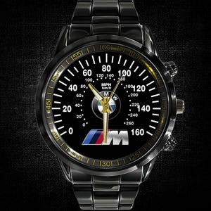 May include: Black wristwatch with a speedometer-style face, featuring white and gold accents. The watch face displays the BMW logo and MPH/km/h markings. The watch has a black metal band and a black bezel.