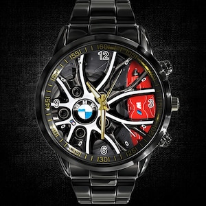 May include: Black wristwatch with a detailed face featuring a BMW wheel design. The watch has a black metal band and a gold-colored bezel. The face includes the BMW logo, white and black accents, and red brake caliper details.