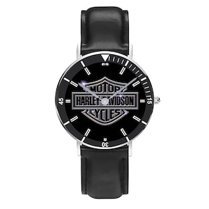 May include: A black wristwatch with a silver-colored bezel and a black leather band. The watch face features the Harley-Davidson logo in silver against a black background. The watch has white hour markers and hands.