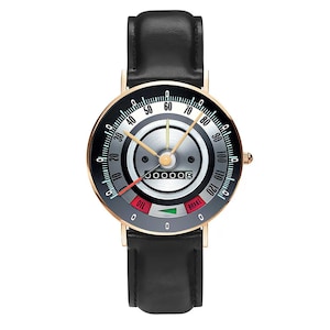May include: A wristwatch with a black leather band and a gold-colored case. The watch face resembles a car speedometer, with a black and silver dial, gold hands, and the word "BRAKE".