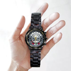 May include: A black wristwatch with a metallic band and a detailed face. The watch face features a speedometer-like design with multiple dials and indicators. The watch is held in a hand, showcasing its design and style.