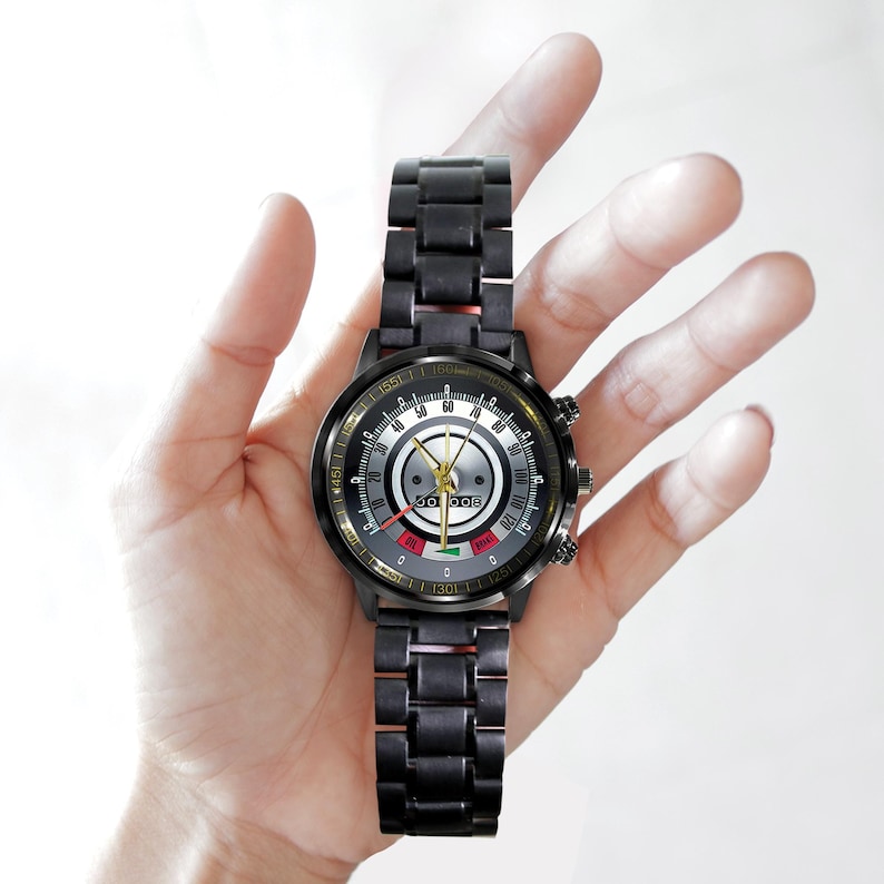 May include: A black wristwatch with a metallic band and a detailed face. The watch face features a speedometer-like design with multiple dials and indicators. The watch is held in a hand, showcasing its design and style.
