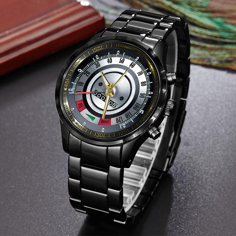 May include: Black wristwatch with a unique dial design. The watch features a black metal band and case, with a dial that includes a speedometer-like display with gold-colored hands and markers. The watch has a sleek, modern aesthetic.