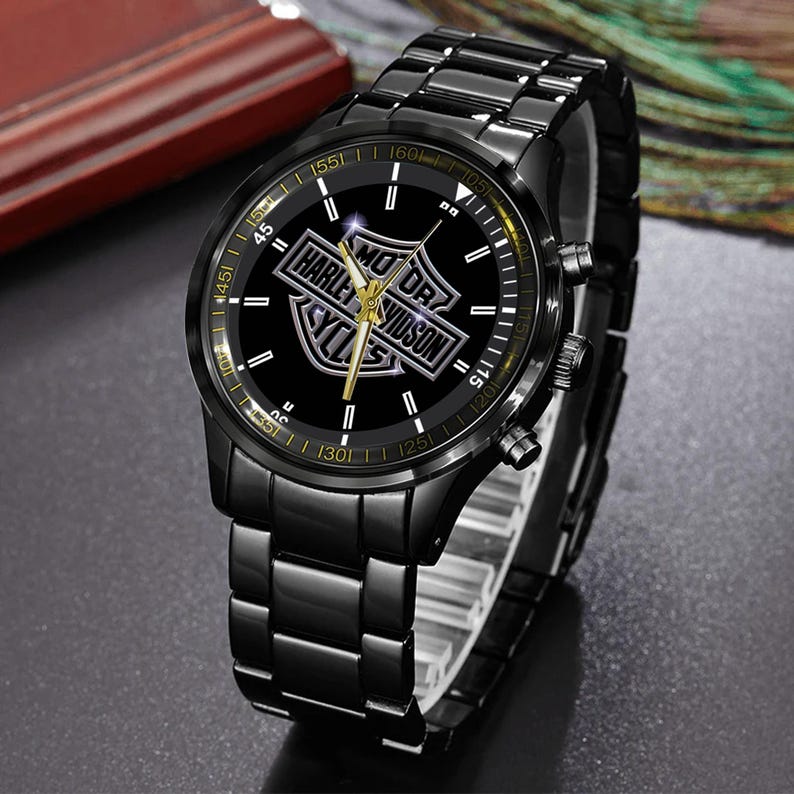 May include: Black wristwatch featuring the Harley-Davidson logo on a black dial with gold accents. The watch has a black metal band and a chronograph design. The watch face displays the Harley-Davidson logo in silver.