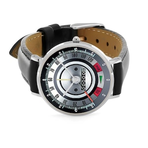 May include: A silver-toned wristwatch with a black leather band. The watch face features a speedometer-style design with a black, white, and gray color scheme. The watch has a red needle and the word "BRAKE" is visible.