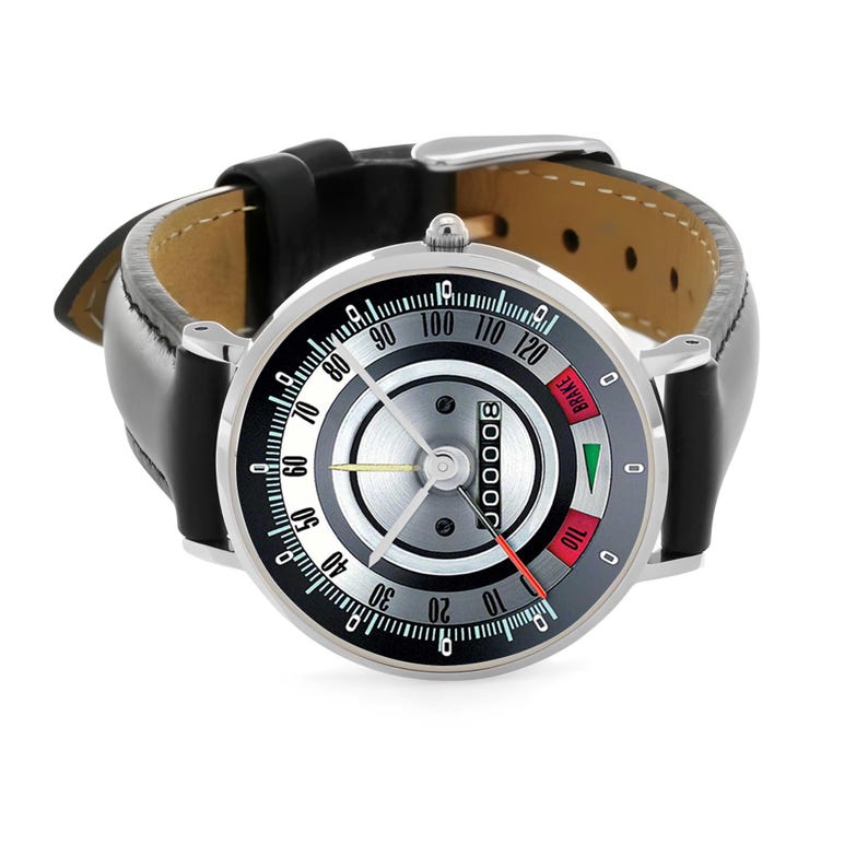May include: A silver-toned wristwatch with a black leather band. The watch face features a speedometer-style design with a black, white, and gray color scheme. The watch has a red needle and the word "BRAKE" is visible.
