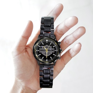May include: Black wristwatch with a metal band. The watch face features the Harley-Davidson logo in gold and white. The watch has a black bezel and a black metal band.