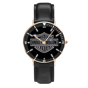 May include: A black watch with a gold-colored bezel and a black leather band. The watch face features the Harley-Davidson logo in silver against a black background. The watch has white markers and hands.