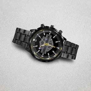 May include: Black wristwatch with a round face featuring the Harley-Davidson logo in gold and white. The watch has a black metal band and a black bezel with gold accents. The watch hands are gold.