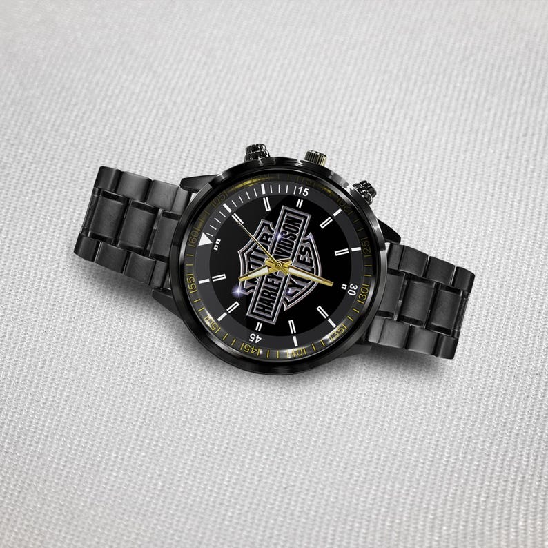 May include: Black wristwatch with a round face featuring the Harley-Davidson logo in gold and white. The watch has a black metal band and a black bezel with gold accents. The watch hands are gold.