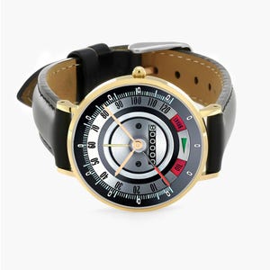 May include: A gold-toned wristwatch with a black leather band. The watch face features a speedometer-style design with a black, silver, and white color scheme. The watch has a red and green indicator and the word "BRAKE".