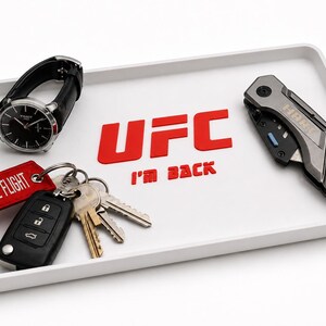 Peut inclure: Un plateau blanc avec le logo UFC et le texte "I'm Back" en rouge. Le plateau contient une montre noire, un jeu de clés, une étiquette rouge "REMOVE BEFORE FLIGHT" et un couteau argenté et noir.