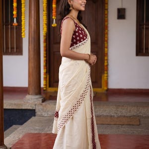 May include: A cream-colored saree featuring a maroon border with a repeating white floral pattern. The sleeveless blouse matches the border, displaying a maroon fabric with a white floral print. The garment drapes elegantly, showcasing the contrast.