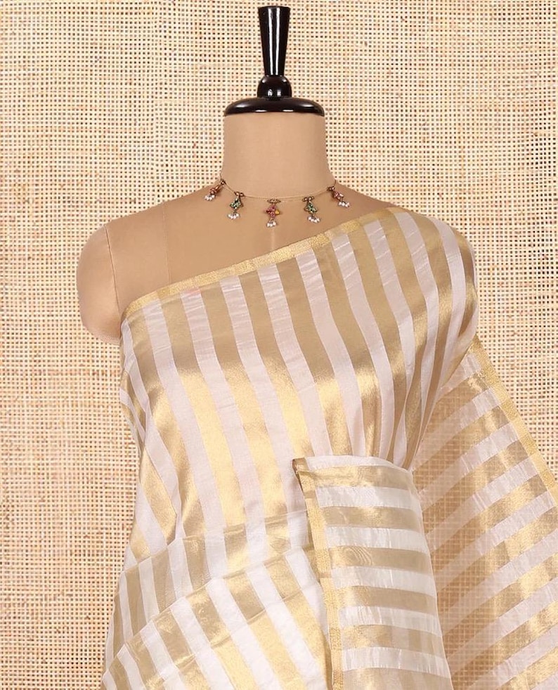 May include: A saree featuring alternating vertical stripes of white and metallic gold fabric. The garment is draped over a mannequin and is paired with a delicate gold-toned choker necklace adorned with small colorful beads and white accents.