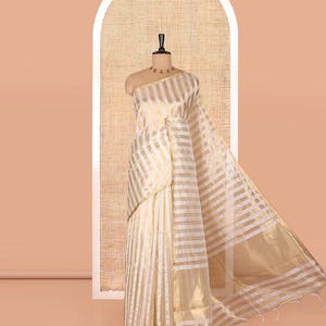 May include: A saree featuring a repeating pattern of horizontal white and metallic gold stripes. The fabric has a semi-sheer quality with a distinct sheen, draped over a mannequin. The garment includes a long, flowing pallu section with fringe.