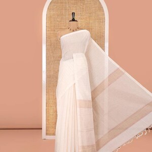 May include: A cream-colored saree draped on a mannequin, featuring a subtle horizontal striped texture throughout the fabric. The garment includes a wide border with light brown horizontal bands and a fringed edge at the end of the pallu.