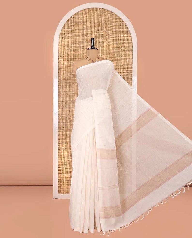 May include: A cream-colored saree draped on a mannequin, featuring a subtle horizontal striped texture throughout the fabric. The garment includes a wide border with light brown horizontal bands and a fringed edge at the end of the pallu.