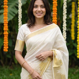 May include: A traditional off-white saree featuring a wide gold zari border along the hem and sleeves. The garment is crafted from a lightweight fabric with a subtle sheen, showcasing a classic design with clean lines and a structured drape.