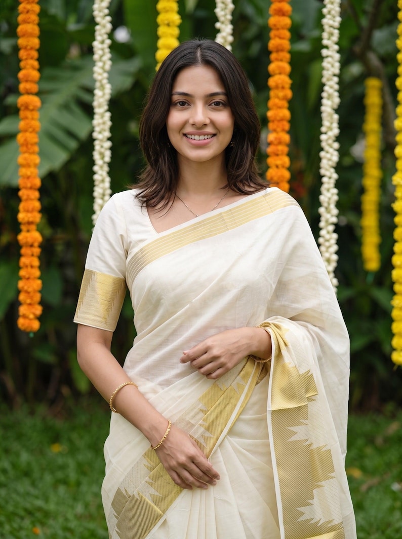May include: A traditional off-white saree featuring a wide gold zari border along the hem and sleeves. The garment is crafted from a lightweight fabric with a subtle sheen, showcasing a classic design with clean lines and a structured drape.