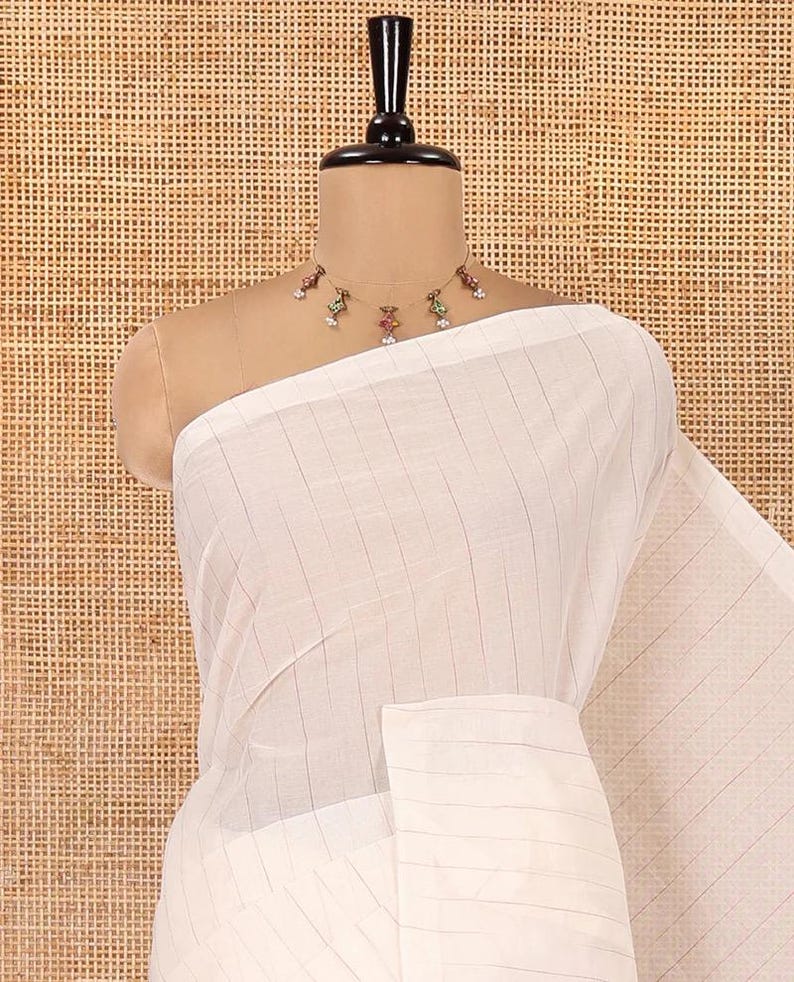 May include: A white saree featuring a subtle vertical pinstripe pattern, draped over a mannequin. The garment is paired with a delicate gold-toned necklace adorned with small, colorful enamel-style floral charms and white beads.