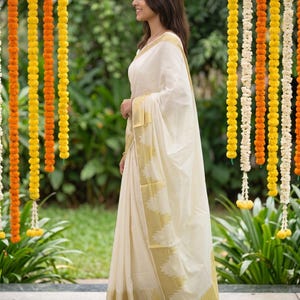 May include: An off-white saree featuring a wide gold border with a triangular serrated pattern along the edge. The fabric is draped in a traditional style, with the pallu flowing over the shoulder, showcasing a smooth, lightweight texture.