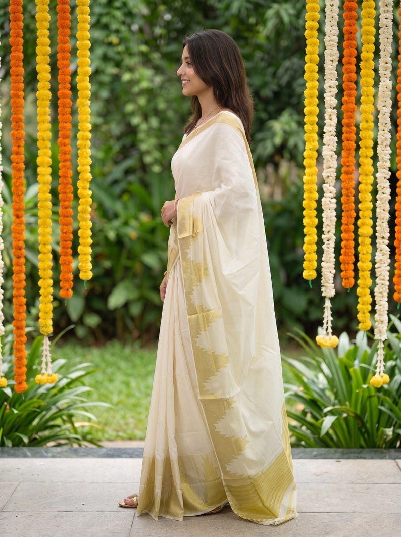 May include: An off-white saree featuring a wide gold border with a triangular serrated pattern along the edge. The fabric is draped in a traditional style, with the pallu flowing over the shoulder, showcasing a smooth, lightweight texture.