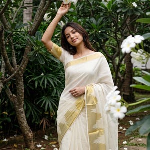 May include: A cream-colored saree featuring a wide gold border with a geometric pattern along the hem and sleeves. The garment is made of a lightweight fabric and is draped over the shoulder, showcasing a clean, solid-colored bodice and skirt.