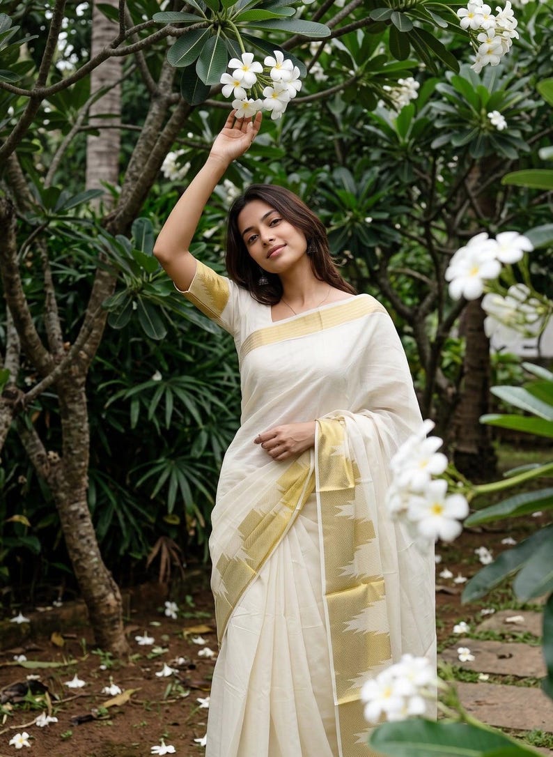 May include: A cream-colored saree featuring a wide gold border with a geometric pattern along the hem and sleeves. The garment is made of a lightweight fabric and is draped over the shoulder, showcasing a clean, solid-colored bodice and skirt.