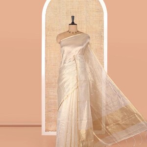 May include: A cream-colored saree featuring a subtle horizontal striped pattern throughout the fabric. The garment includes a wide, metallic gold border at the hem and a matching gold trim along the edges, displayed on a mannequin form.