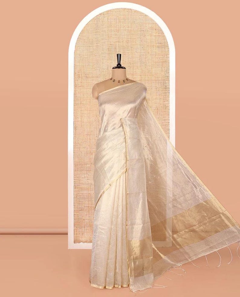 May include: A cream-colored saree featuring a subtle horizontal striped pattern throughout the fabric. The garment includes a wide, metallic gold border at the hem and a matching gold trim along the edges, displayed on a mannequin form.