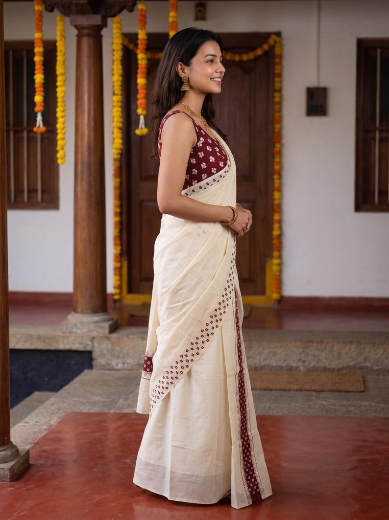 May include: A cream-colored saree featuring a maroon border with a repeating white floral pattern. The sleeveless blouse matches the border, displaying a maroon fabric with a white floral print. The garment drapes elegantly, showcasing the contrast.