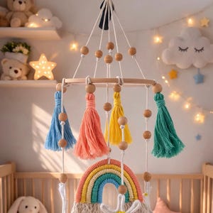 May include: A handmade baby mobile featuring a rainbow, tassels, and wooden beads. The mobile has a wooden ring and is suspended from a white cord. The tassels are blue, coral, yellow, and green. The rainbow has multiple colors.