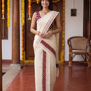 May include: A cream-colored saree featuring a deep maroon border with a repeating geometric pattern. The ensemble includes a sleeveless maroon blouse with a small white print, a gold necklace, and gold bangles, creating a coordinated look.