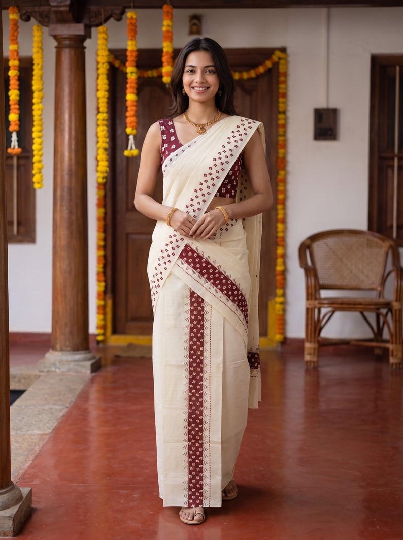May include: A cream-colored saree featuring a deep maroon border with a repeating geometric pattern. The ensemble includes a sleeveless maroon blouse with a small white print, a gold necklace, and gold bangles, creating a coordinated look.