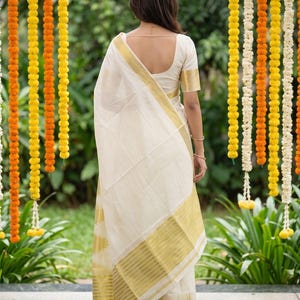 May include: A cream-colored saree featuring a wide gold border with horizontal stripes and a matching short-sleeved blouse. The garment is draped over the shoulder, showcasing the metallic gold detailing along the hem and the fabric's texture.