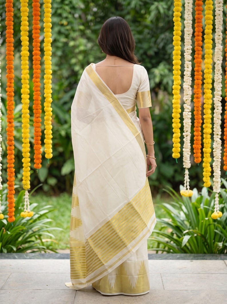 May include: A cream-colored saree featuring a wide gold border with horizontal stripes and a matching short-sleeved blouse. The garment is draped over the shoulder, showcasing the metallic gold detailing along the hem and the fabric's texture.
