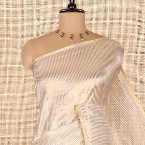 May include: A cream-colored saree featuring a subtle, repeating vertical striped pattern throughout the fabric. The garment is draped on a mannequin and is paired with a delicate gold-toned choker necklace adorned with small, colorful, dangling accents.