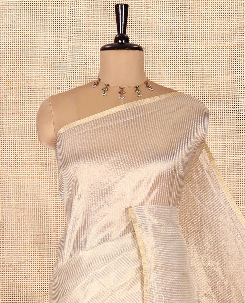 May include: A cream-colored saree featuring a subtle, repeating vertical striped pattern throughout the fabric. The garment is draped on a mannequin and is paired with a delicate gold-toned choker necklace adorned with small, colorful, dangling accents.