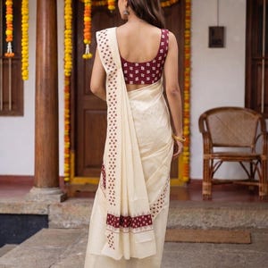 May include: A cream-colored saree featuring a maroon sleeveless blouse with a small white geometric pattern. The saree includes a matching maroon and white patterned border that runs along the edge and drapes over the shoulder.