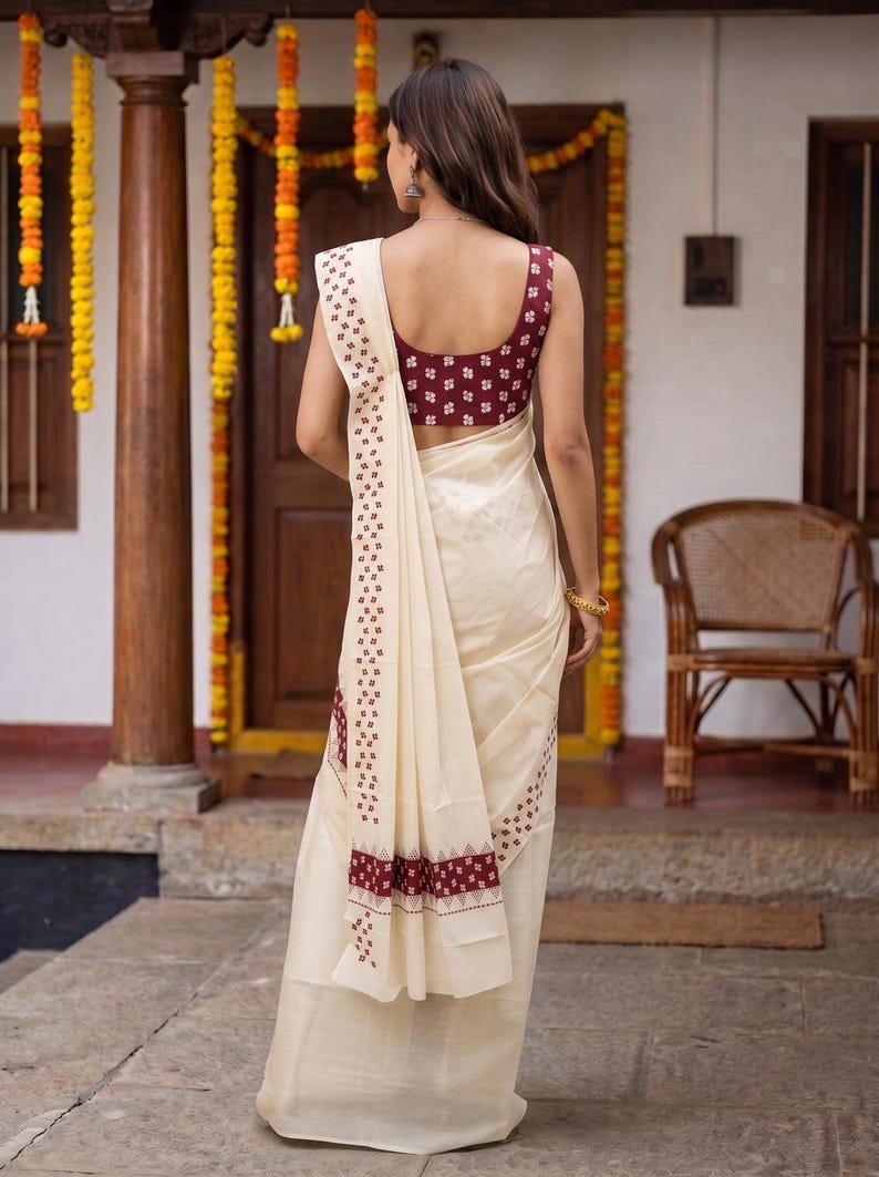 May include: A cream-colored saree featuring a maroon sleeveless blouse with a small white geometric pattern. The saree includes a matching maroon and white patterned border that runs along the edge and drapes over the shoulder.