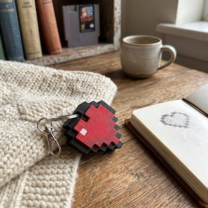 May include: A pixelated red and black keychain in the shape of a video game character. The keychain is attached to a silver clasp. The background includes a mug, a notebook, and a stack of books.
