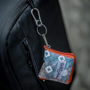 Könnte beinhalten: Kleine, quadratische Tasche mit Batikmuster und orangefarbenem Reißverschluss, befestigt an einem schwarzen Rucksack mit einem silbernen Karabinerhaken. Die Tasche hat ein weißes Etikett mit der Aufschrift "BeBa Collection". Die Wörter "Handicraft 100% Handmade" und "Beba Collection" sind ebenfalls sichtbar.