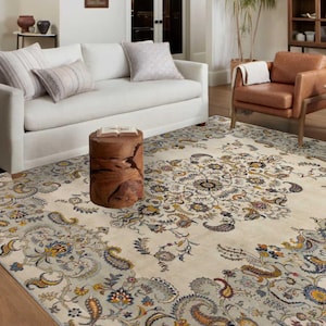 May include: A large, cream-colored area rug with an intricate floral and paisley design. The rug features a central medallion and borders with blue, yellow, and gray accents. The rug is placed in a living room setting with a white sofa and brown leather chairs.