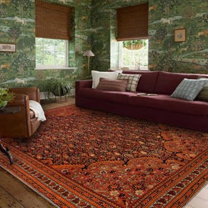 May include: A living room scene showcasing a richly patterned rug with a red and orange floral design. The room includes a burgundy sofa, cushions, and green wallpaper depicting a pastoral scene, creating a warm and inviting atmosphere.